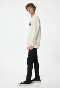 Sudadera crema con letras oscuras, pantalones ajustados negros y zapatillas altas negras con suelas beige, contra un fondo liso.