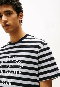 Czarny i szary pasek z krótkim rękawem, z białym napisem "TOMMY JEANS N.Y.C. UNIVERSITY CLUB" z przodu oraz logo na rękawie.