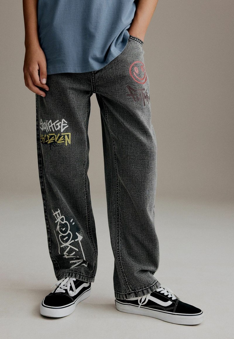 Personne portant un jean gris ample avec des motifs de style graffiti et des baskets noir et blanc, debout avec une main dans la poche.