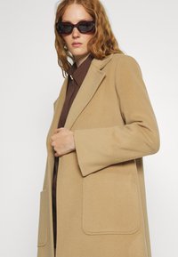 Gürtelter Woll-Mischmantel in hellem Beige mit Spitzrevers, zwei schlichten Fronttaschen und einer weichen Textur, getragen über einem braunen Hemd.