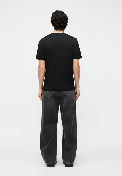 Άτομο με σγουρά, σκούρα μαλλιά στέκεται με την πλάτη, φορώντας μια απλή μαύρη μπλούζα και φαρδιά σκούρα γκρι τζιν μπροστά σε φωτεινό φόντο.