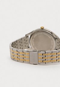 Hopeakultainen ruostumattomasta teräksestä valmistettu kello, jossa on linkkimäinen ranneke, pyöreä kotelo, kullattu kruunu ja kaiverrettu takapohja vedenkestävyydelle.