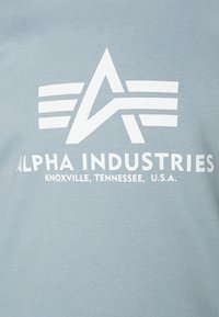 T-shirt azzurro chiaro con un grande logo grafico bianco caratterizzato da un design angolare e testo che riporta "Alpha Industries, Knoxville, Tennessee, U.S.A."