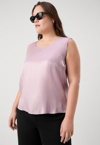Lys rosa ermeløs topp laget av glatt stoff, med rund hals og avslappet passform, sammen med svarte bukser og kantete solbriller.