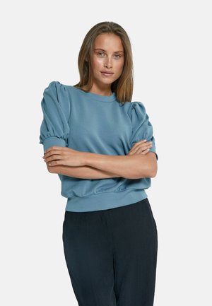 Light blue sweatshirt with puffed sleeves, round neckline, and gathered hem. Paired with dark pants, showcasing a relaxed fit design.