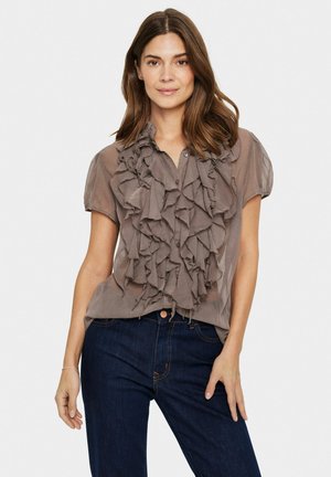 Camicia beige chiaro a maniche corte con multiple rouches che scendono davanti, chiusura a bottoni e tessuto morbido e trasparente. Indossata con jeans scuri.