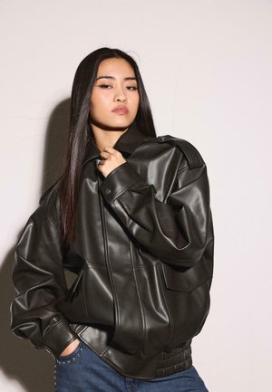 Žena s dugom tamnom kosom nosi crnu preveliku kožnu jaknu i plave traperice, pozirajući s jednom rukom blizu ovratnika.