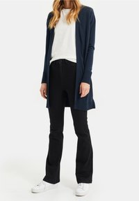 Mörkblå stickad cardigan med ribbad textur och öppen framsida, bärs över en vit T-shirt och svarta utsvängda jeans, kombinerad med vita sneakers.