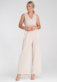 Beżowy kombinezon z dekoltem w kształcie litery V, szerokimi nogawkami, marszczonym w talii z paskiem i lekkim materiałem. Połączony z sandałami na obcasie i dodatkami.