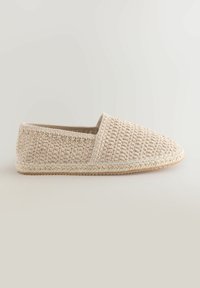 Zapato slip-on de tejido beige con puntera redondeada, parte superior texturizada y suela de yute trenzado. Diseño plano, detalle de costura minimalista alrededor de los bordes.