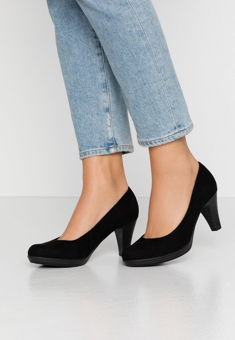 Des pieds chaussés de chaussures à talons mi-hauts en daim noir, associés à un jean cropped bleu clair, posés sur un sol blanc lisse.