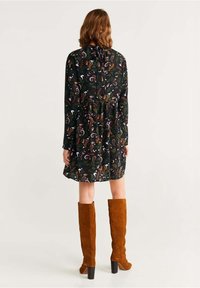 Schwarzes Kleid mit mehrfarbigem Paisleymuster, raffiniert in der Taille, lange Ärmel, kombiniert mit kniehohen braunen Wildlederstiefeln und einem blockigen Absatz.