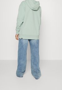 Felpa verde chiaro con tasca a marsupio e orlo a coste abbinata a jeans blu chiari e larghi. Presenta un'etichetta con logo sui jeans.