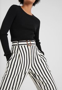 Top lungo a maniche nere a costine abbinato a pantaloni bianchi a vita alta con audaci strisce verticali nere e un dettaglio di fibbia marrone.