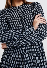 Kobieta w czarnej sukience z długim rękawem, ozdobionej jasnoniebieskimi owalnymi wzorami, z guzikami z przodu, z założonymi rękami.