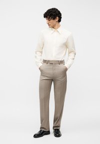 Άνδρας που στέκεται με τα χέρια στις τσέπες, φορώντας κρεμ πουκάμισο με κουμπιά, μπεζ ριγέ παντελόνι και μαύρα επαγγελματικά παπούτσια, κοιτάζοντας προς τα πλάγια.