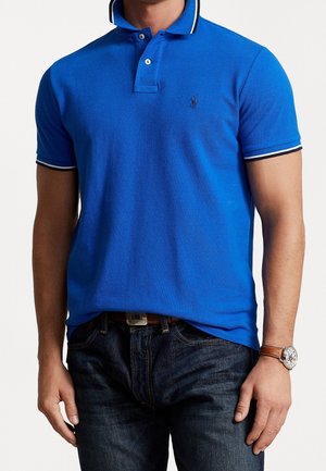 Polo bleu en maille, doté d'un col classique et d'une patte à deux boutons, avec un bord blanc sur les manches. Porté avec un jean en denim.