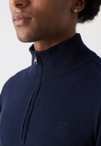 Suéter azul marino con cremallera y cuello alto, textura de canalé y detalle de logo bordado. Primer plano en la zona del cuello y el hombro.
