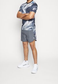 Camiseta atlética gris con diseño abstracto en blanco, combinada con pantalones cortos grises a juego. Zapatillas blancas con rayas negras completan el conjunto.