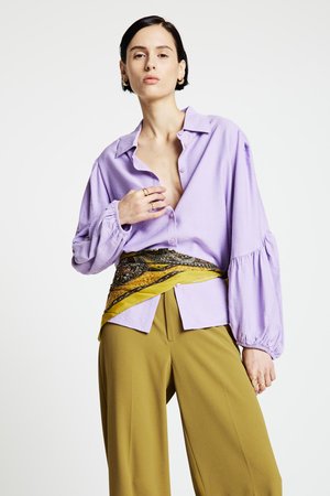 Lavendelfarbenes Hemd mit Knöpfen und Puffärmeln, kombiniert mit einem gelben, gemusterten Schal und hoch taillierter olivgrüner Hose.