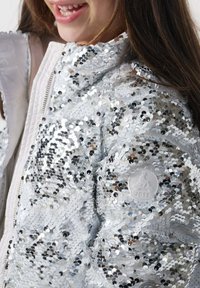 Chaqueta bomber de lentejuelas plateadas con una textura brillante, que presenta un cierre de cremallera y un parche de logo redondo en la manga. Diseño plateado y reflectante.