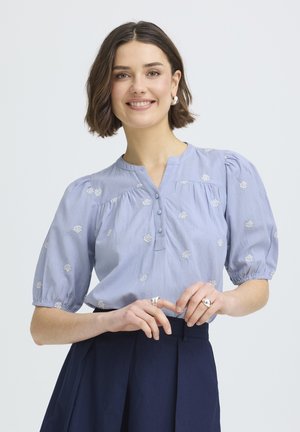 Frau mit kurzen braunen Haaren, die eine hellblaue Bluse mit Puffärmeln und eine dunkle, hoch taillierte Hose trägt, lächelt und hält die Hände gefaltet.