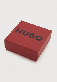 Κόκκινο ορθογώνιο κουτί με ματ φινίρισμα, με το μαύρο τυπωμένο κείμενο «HUGO» στην πάνω επιφάνεια. Κλειστό σχέδιο, χωρίς ορατό μηχανισμό.