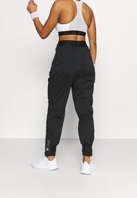 Czarne spodnie przeciwwiatrowe z elastycznymi mankietami, bocznymi kieszeniami i naszywką z logo. W zestawie z białymi butami sportowymi i białym biustonoszem sportowym.