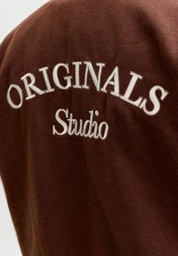 Brunt stoff med hvit brodering som staver "ORIGINALS Studio" i en buet utforming, og fremhever tekstur og håndverk.