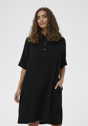 Kobieta z kręconymi włosami ubrana w luźną czarną sukienkę do kolan z krótkimi rękawami, zapinany kołnierzyk na guziki, z ręką w bocznej kieszeni.