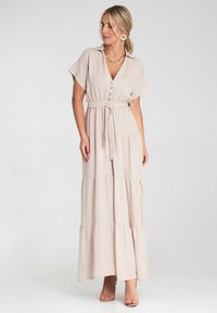 Długa, beżowa sukienka maxi z krótkimi rękawami, zapinana na guziki dekolt w serek, podkreślona talia i wielopoziomowy design z falbanami. Gładki materiał, minimalistyczny styl.