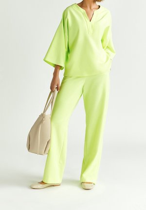 Woman wearing a loose neon yellow top and matching pants, holding a beige handbag in one hand with beige flats on white background.