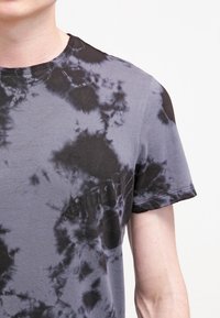 Šedé tričko s krátkým rukávem v motivu tie-dye. Má měkkou texturu, nepravidelné černé vzory a klasický kulatý výstřih.