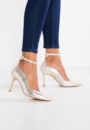 Vrouw draagt glanzende zilveren enkellaarzen met hoge hakken, gecombineerd met donkerblauwe skinny jeans, staand op een witte vloer tegen een effen achtergrond.