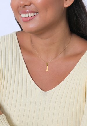 Goldkette mit einem Anhänger in Form eines kleinen "j". Das Model trägt ein hellgelbes geripptes Oberteil, das das Design und die Farbe der Kette betont.