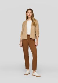 Beiger, flauschiger Cardigan über einem weißen Hemd, kombiniert mit braunen taillierten Hosen und weißen Sneakers mit braunen Sohlen. Minimalistisches Design, weiche Textur.