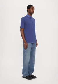 Polo shirt in dark blue cotton with a collar, short sleeves, and three-button placket, paired with light blue wide-leg jeans and black shoes.