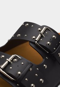 Μαύρα λουριά σανδαλιών με ασημένιες αγκράφες και στρογγυλές μεταλλικές καρφίτσες σε ταμπά εσωτερική σόλα, κοντινή όψη.