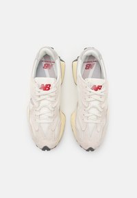 Weiße Sneakers aus Wildleder und Mesh mit roten Akzenten, einer kräftigen gelben Sohle und strukturierten Überzügen. Schnürsenkel und Markenlogo auf der Zunge sichtbar.