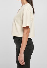 Donna in piedi di lato, indossa una maglietta corta a maniche corte color crema e jeans neri a vita alta, contro uno sfondo neutro.