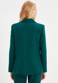 Blazer teal con un design strutturato, caratterizzato da un colletto a risvolto e un tessuto liscio. Le cuciture visibili e il taglio su misura mettono in risalto il retro.