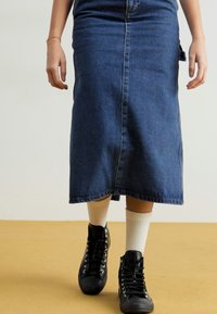 Jupe en denim bleu foncé, de longueur mi-cuisse avec une fente à l'avant. Associée à des baskets montantes noires et des chaussettes blanches. Design simple, sans motifs visibles.