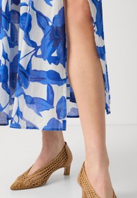 Robe mi-longue avec un motif floral bleu sur un tissu blanc ; associée à des chaussures à talons tressées beiges avec un bout pointu et un talon bas.