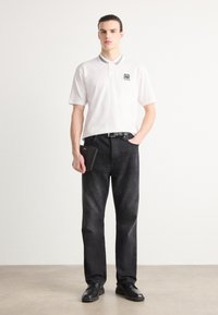 Witte poloshirt met een ribgebreide kraag en logo, gecombineerd met donkere spijkerbroeken en zwarte leren schoenen. Houdt een zwarte portemonnee vast. Eenvoudige stijl.