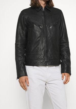 Homme portant une veste en cuir noire avec fermeture éclair à l'avant et poches zippées sur la poitrine, associée à un pantalon blanc, debout devant un fond uni.