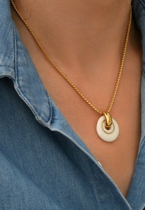 Collier en chaîne en or avec un pendentif circulaire blanc et un petit anneau en or, porté sur le col d'une chemise en denim.