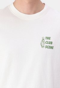 Hvid bomulds t-shirt med en grøn grafik af en hånd, der holder et golfkølle, med teksten "THE CLUB SCENE" i grøn skrift.
