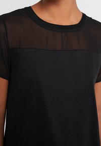 Camiseta negra con un panel transparente en la parte superior, escote redondo, mangas cortas y textura suave. Presenta costuras sutiles.