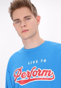 Kék pamut pulóver, amelynek elején feltűnő fehér és piros szöveg olvasható: "LIVE TO Perform", kerek nyakkivágással.
