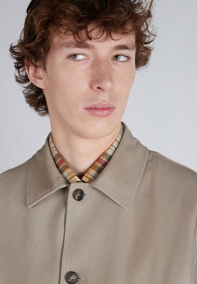 Cappotto in suede beige chiaro con colletto aperto, con una camicia a quadri multicolore sotto. Due bottoni scuri decorano la parte anteriore.